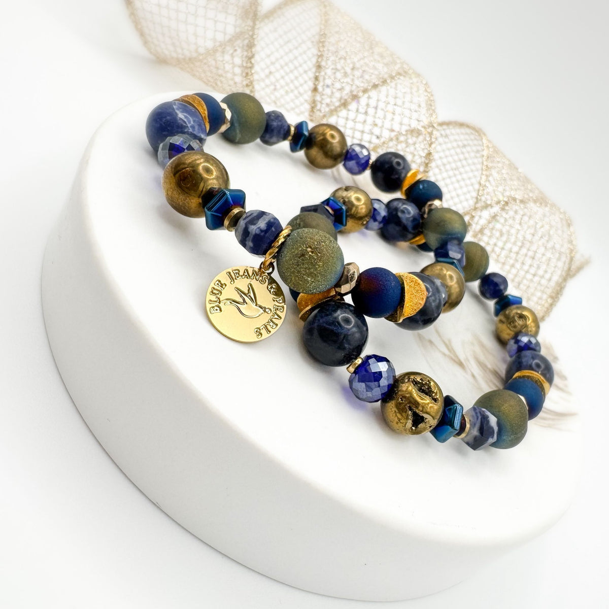 Between Stars Subdued Sparkle handmade sodalite and geode druzy bracelet pair displayed on white pedestal with gold mesh ribbon, featuring olive green and gold druzy, blue hematite and faceted glass with gold Blue Jeans and Pearls charm, one of a kind jewelry Saskatchewan