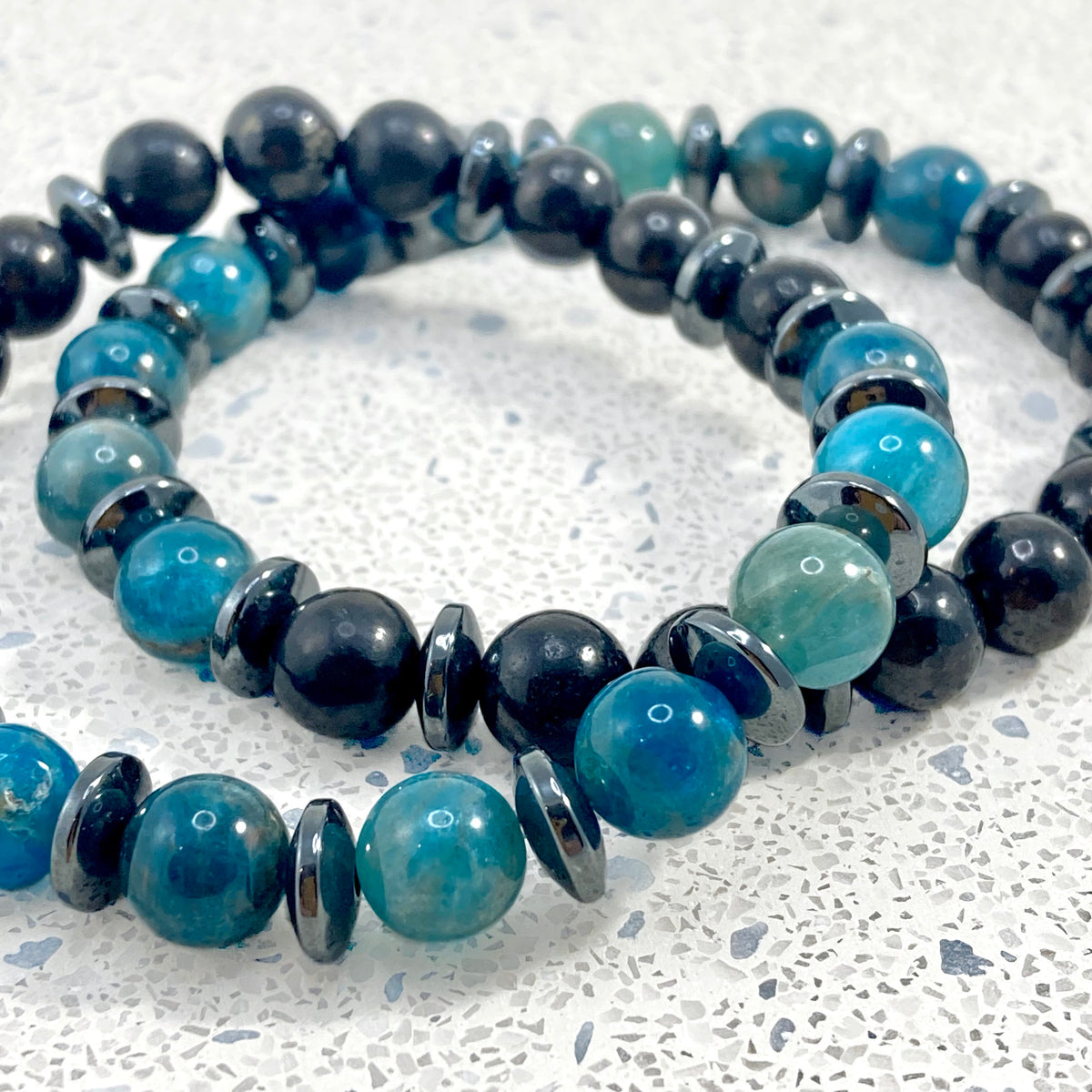 Men's Apatite, Shungite and Hematite handmade bracelet pair close up showing natural blue and teal apatite variations, polished black shungite and glossy hematite spacers on terrazzo, one of a kind men's crystal jewelry by Blue Jeans and Pearls Saskatchewan