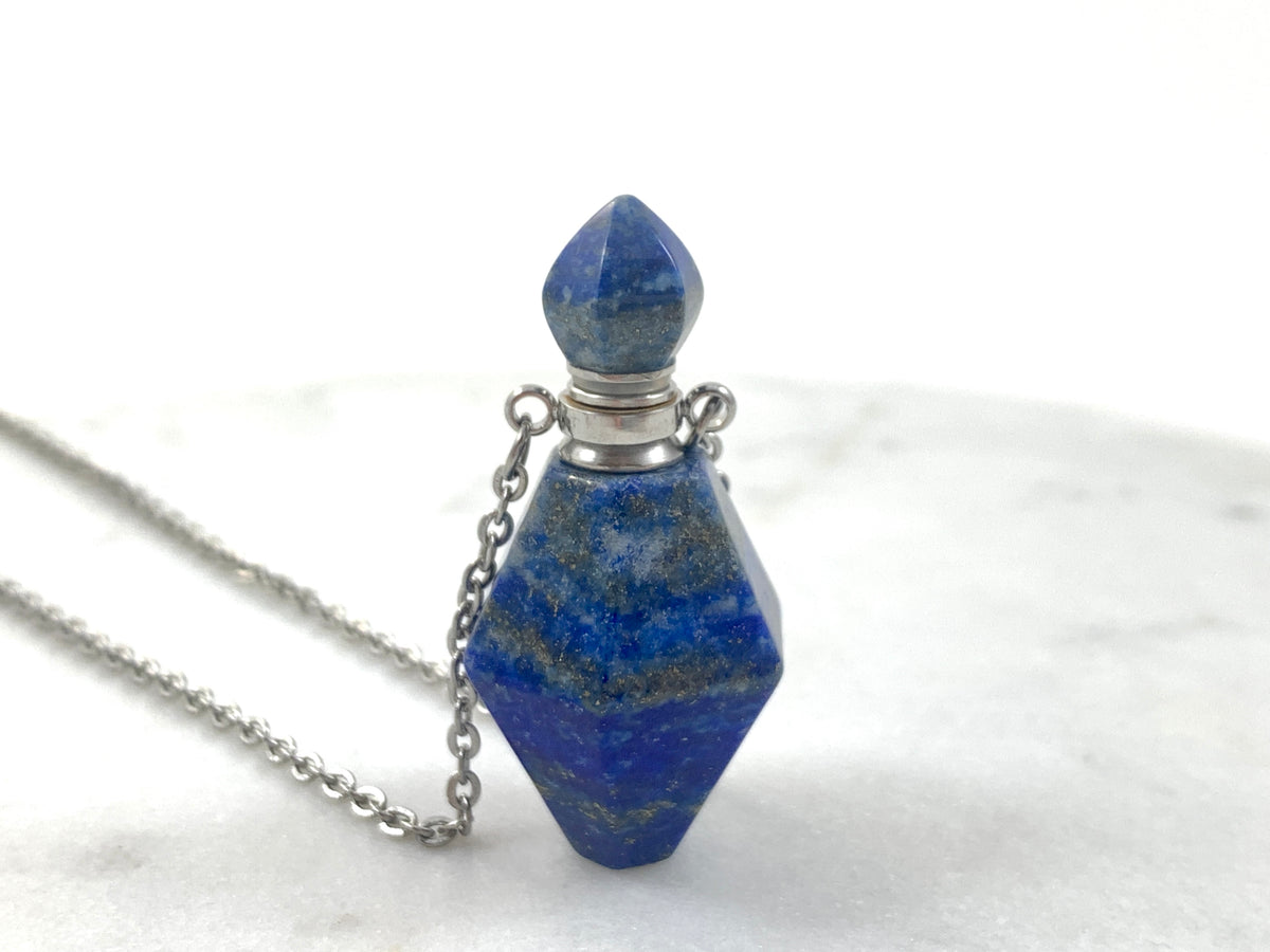 Silver Lapis Lazuli Love Potion Bottle Necklace with a faceted deep blue lapis lazuli crystal bottle pendant flecked with gold pyrite, silver tone cap and stopper on a silver chain, displayed on white marble, essential oil and perfume vial necklace by Blue Jeans and Pearls Saskatchewan