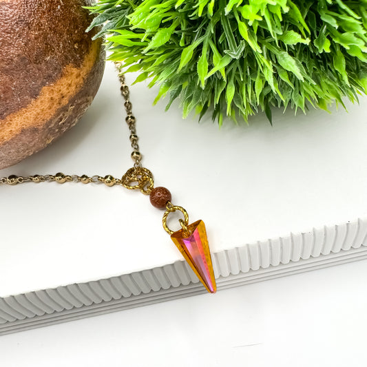 Astral Glow Necklace with an 18mm orange and pink Astral Pink crystal spike pendant and sparkling goldstone bead on a gold-plated beaded chain, styled on a white ribbed stand with greenery and a stone sphere, by Blue Jeans and Pearls Saskatchewan