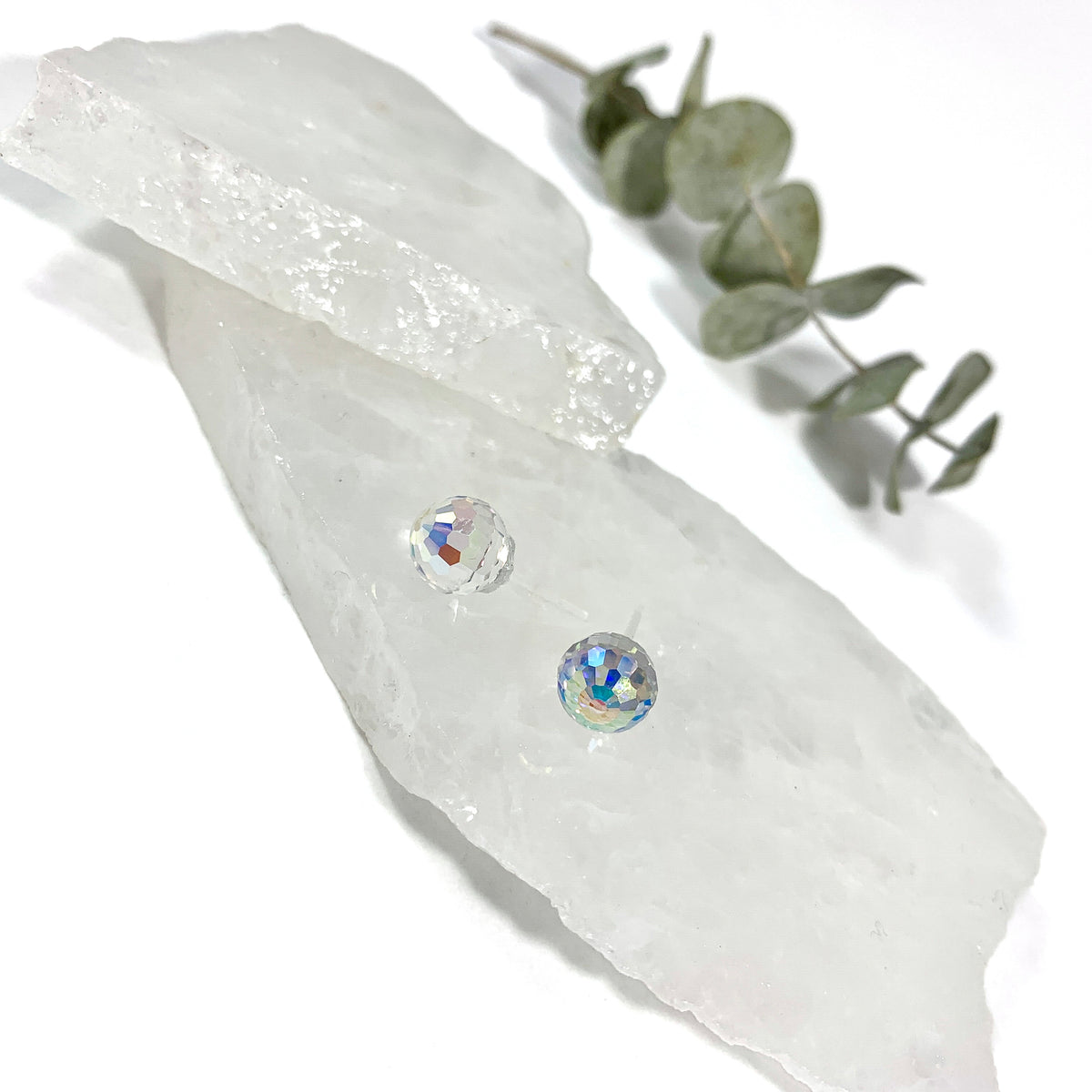 Aurora Borealis disco stud earrings overhead view with 8mm faceted Austrian crystal and rainbow iridescent sparkle on stacked raw white crystal slabs with eucalyptus, minimalist stud earrings for sensitive ears by Blue Jeans and Pearls Saskatchewan