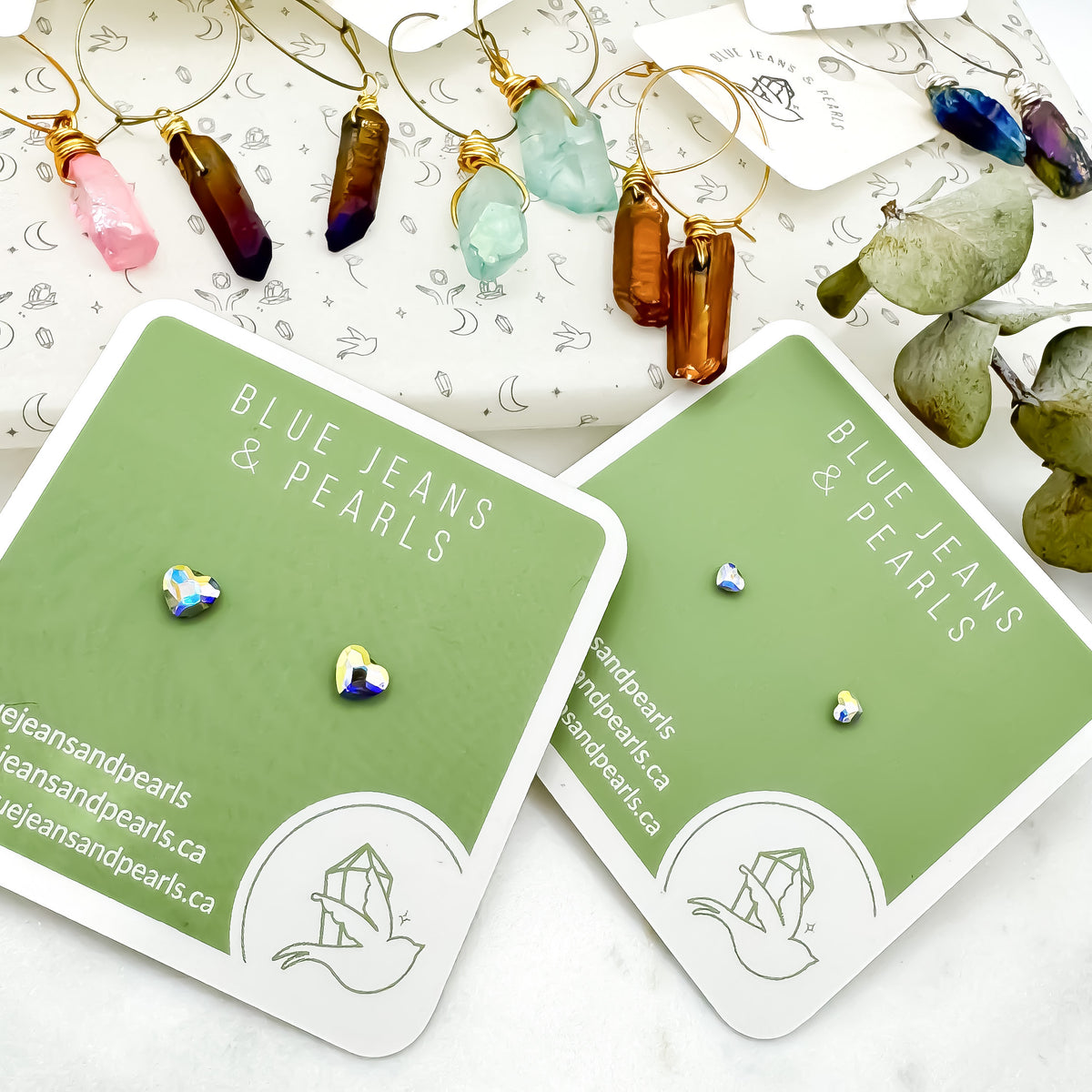 Aurora Borealis heart stud earrings in two sizes on sage green Blue Jeans and Pearls cards, with gold wire-wrapped colourful crystal point hoop earrings on branded tissue paper and eucalyptus, Austrian crystal studs for sensitive ears, Saskatchewan