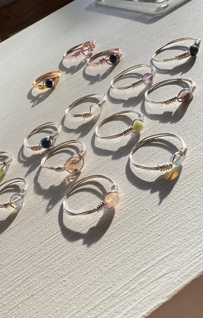 Assortment of Beaded Gemstone Wire Wrap Rings in silver, gold and rose gold finish copper wire laid out in warm sunlight on a white wood surface, showing a variety of black, purple, pink, blue, yellow and iridescent beads, handmade rings by Blue Jeans and Pearls Saskatchewan