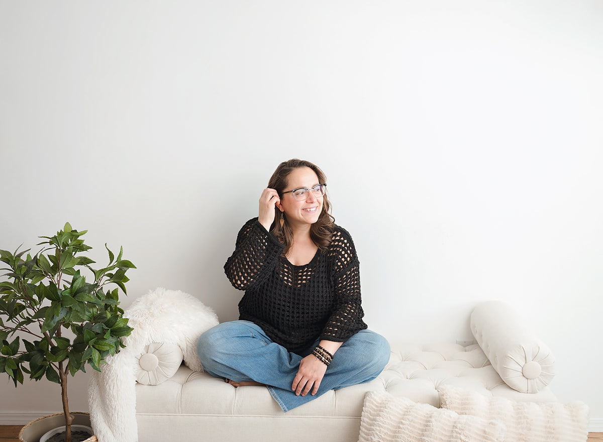 Blue Jeans and Pearls founder Megan Rae Knowles laughing as she sits cross-legged on a white tufted bench in blue jeans and a black crochet sweater, wearing a stack of her handmade bracelets