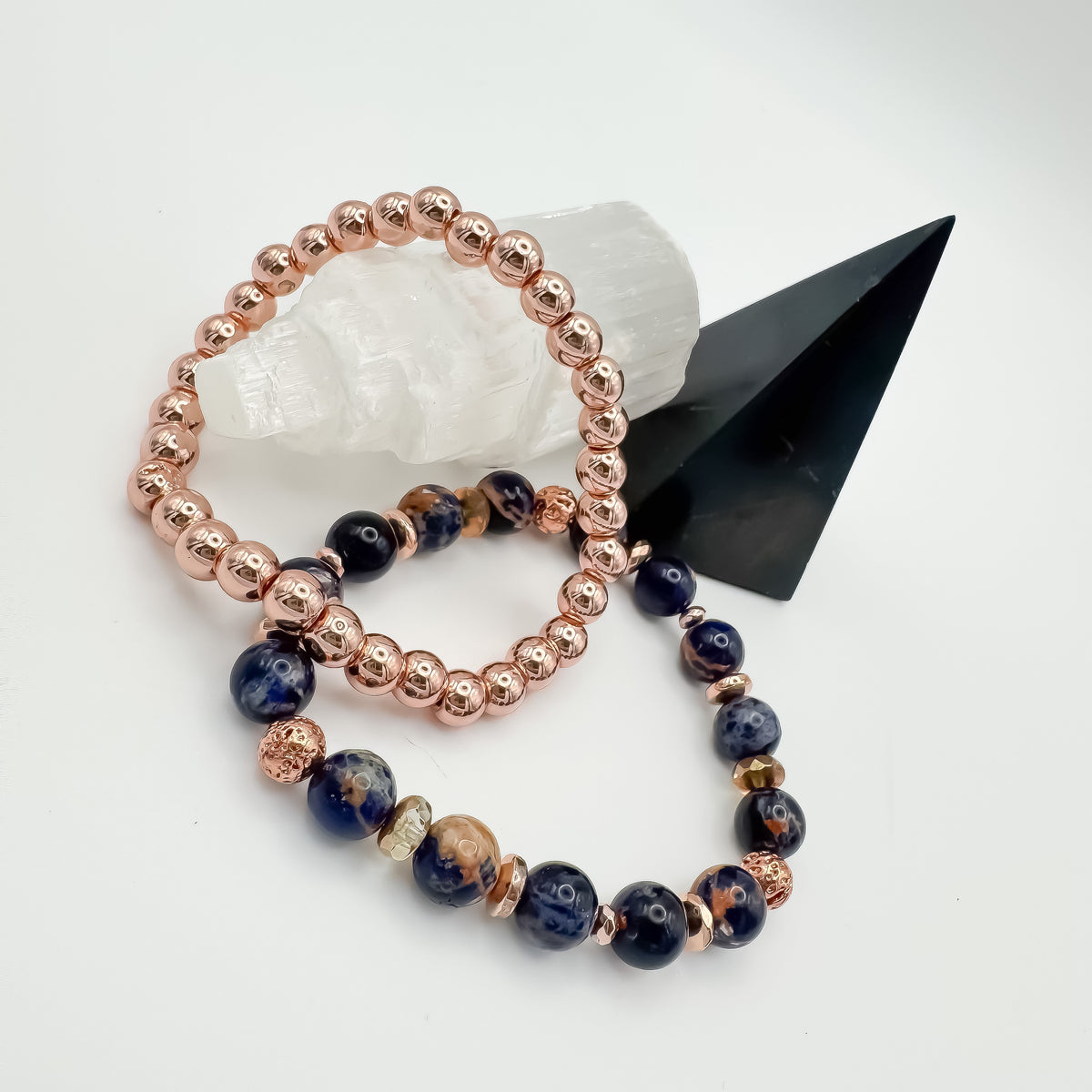 Brazilian sodalite and rose gold lava stone handmade bracelet duo showing full rose gold and sodalite dominant variations on selenite crystal tower with black geometric display, stackable healing jewelry by Blue Jeans and Pearls Saskatchewan
