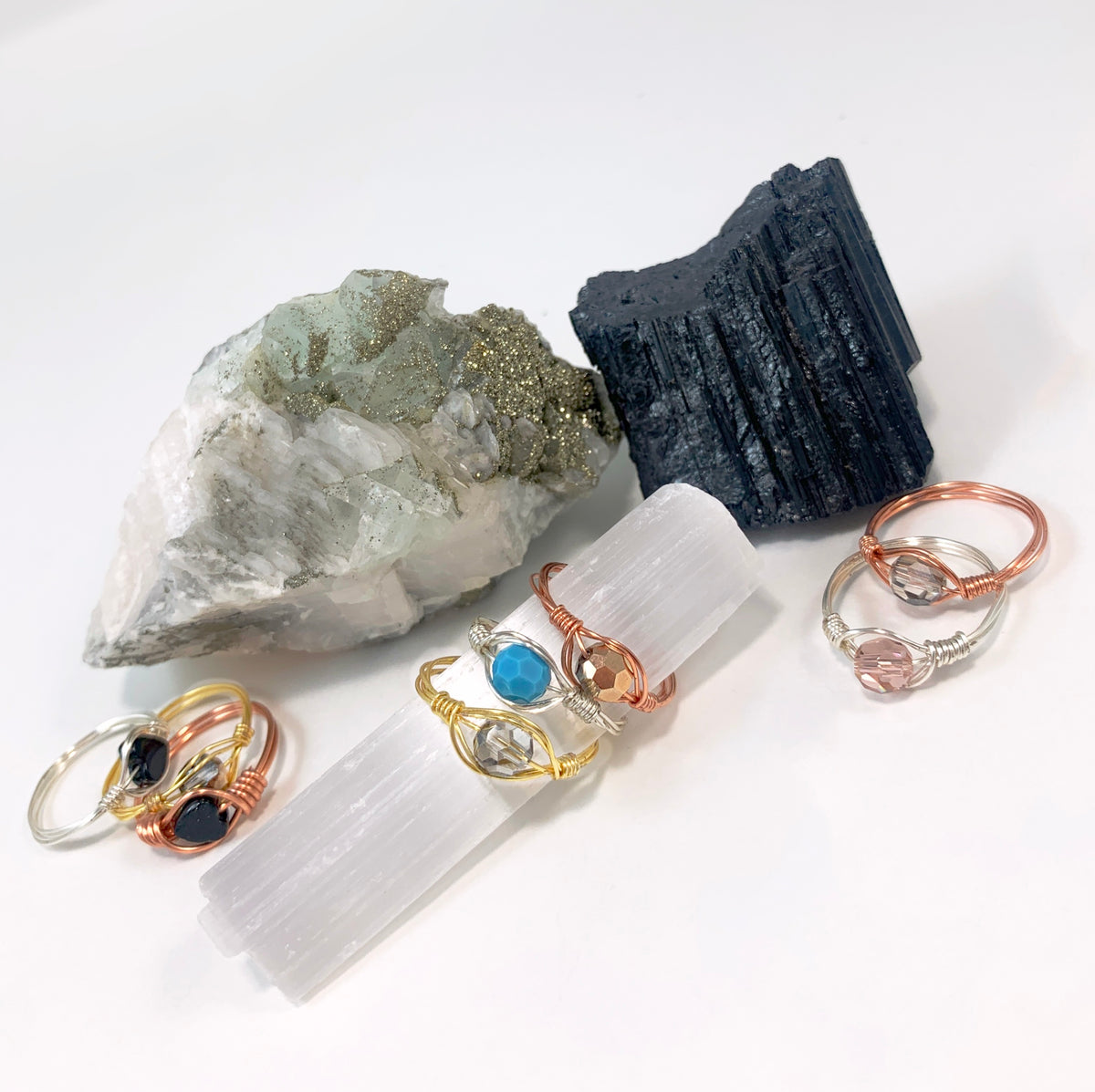 Collection of handmade wire-wrapped gemstone and crystal rings in gold, rose gold and silver finish copper wire, styled on a selenite wand with raw black tourmaline and pyrite crystals, by Blue Jeans and Pearls Saskatchewan