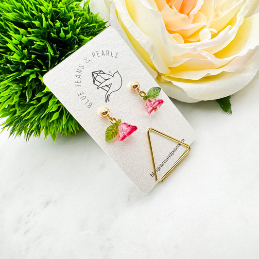 Forever Flowers Roses are Red earrings with pink glass bell flower charms and green glass leaves on 14K gold ball posts, displayed on Blue Jeans and Pearls earring card with gold triangle stand beside cream rose and greenery, handmade floral earrings Saskatchewan