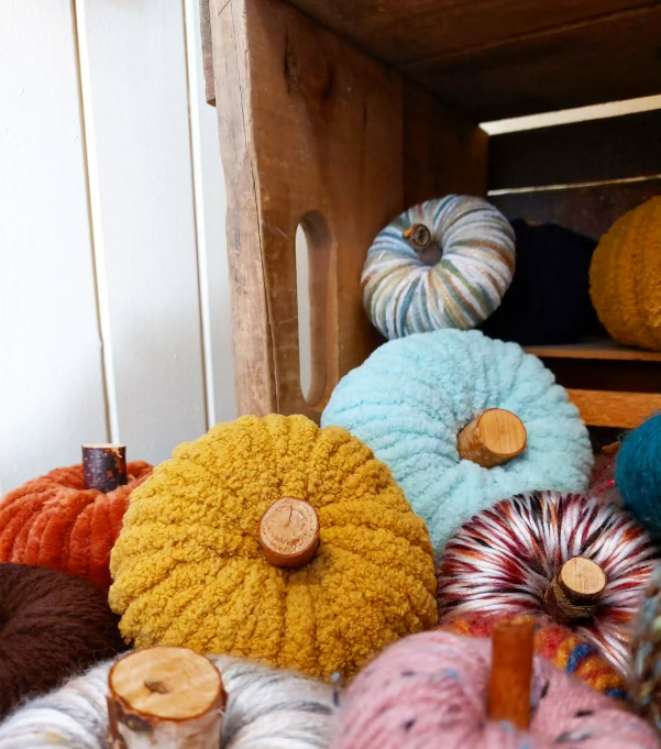 Handmade yarn pumpkins with natural wood branch stems in mustard, sky blue, burnt orange, pink and variegated yarns, piled in a rustic wooden crate, made at the DIY pumpkin decor workshop in Saskatchewan Beach