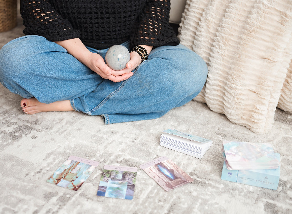 Megan Rae Knowles seated cross-legged in blue jeans holding a speckled crystal sphere, with angel cards spread out on a soft rug in front of her, setting the scene for a one-on-one angel card reading