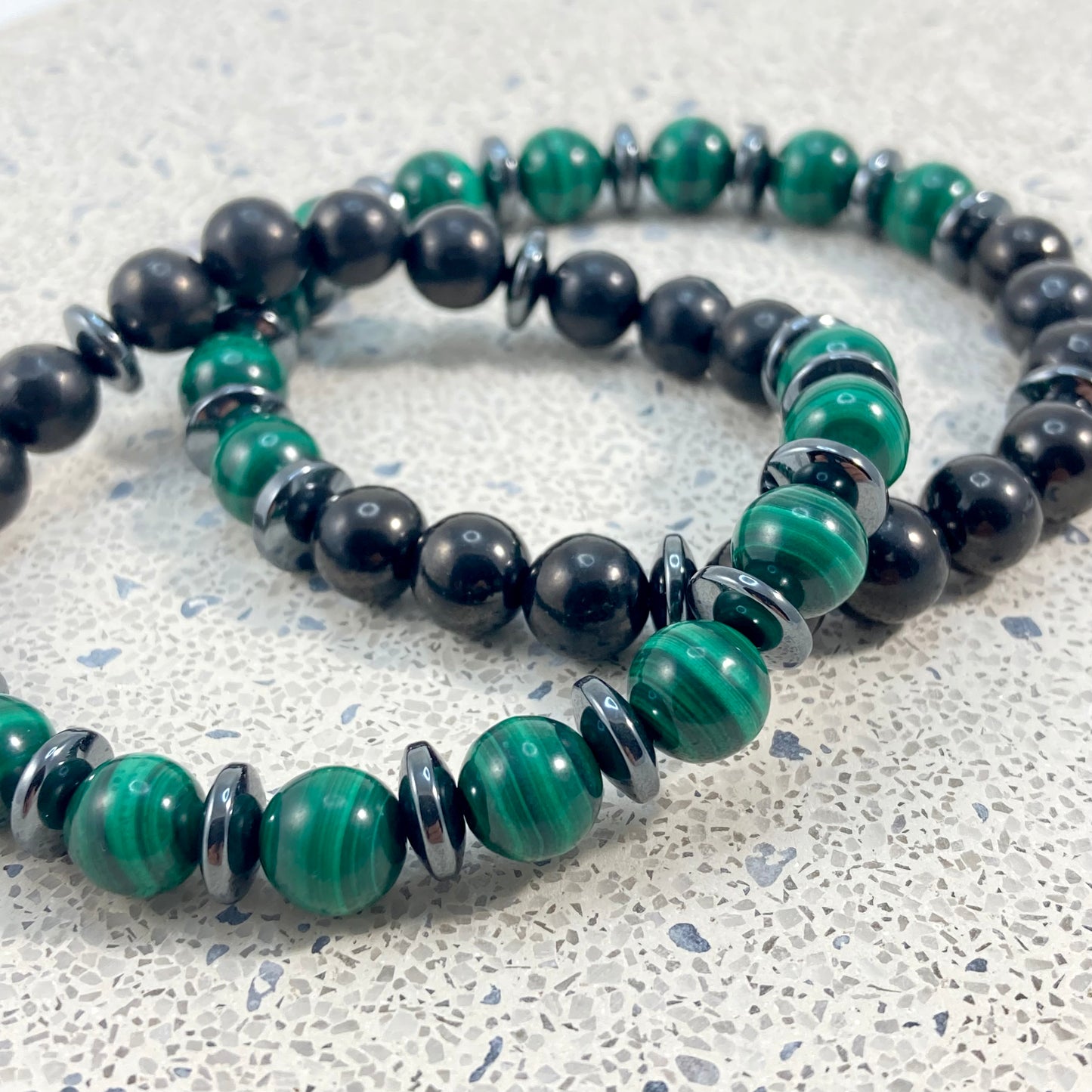 Men's Malachite, Shungite and Hematite handmade bracelet pair close up showing natural green malachite banding, glossy black shungite and polished hematite spacers on terrazzo, one of a kind men's crystal jewelry by Blue Jeans and Pearls Saskatchewan