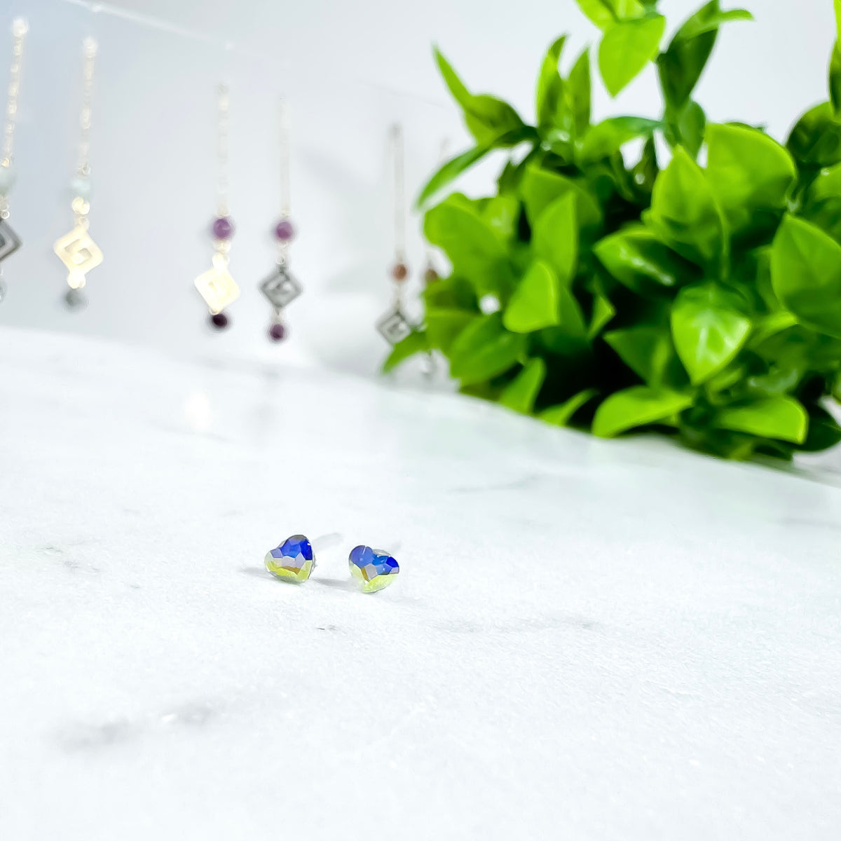 Mini Aurora Borealis heart stud earrings with tiny Austrian crystal hearts showing blue and gold rainbow sparkle on white marble with green plant, dainty stud earrings for sensitive ears by Blue Jeans and Pearls Saskatchewan