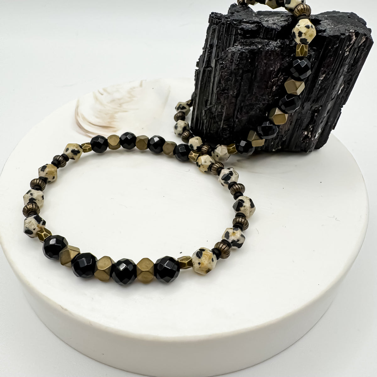Parallel Path handmade bracelet with 6mm dalmatian jasper, faceted black onyx and antique brass tone geometric beads draped on raw black tourmaline and white pedestal with seashell, grounding gemstone jewelry by Blue Jeans and Pearls Saskatchewan