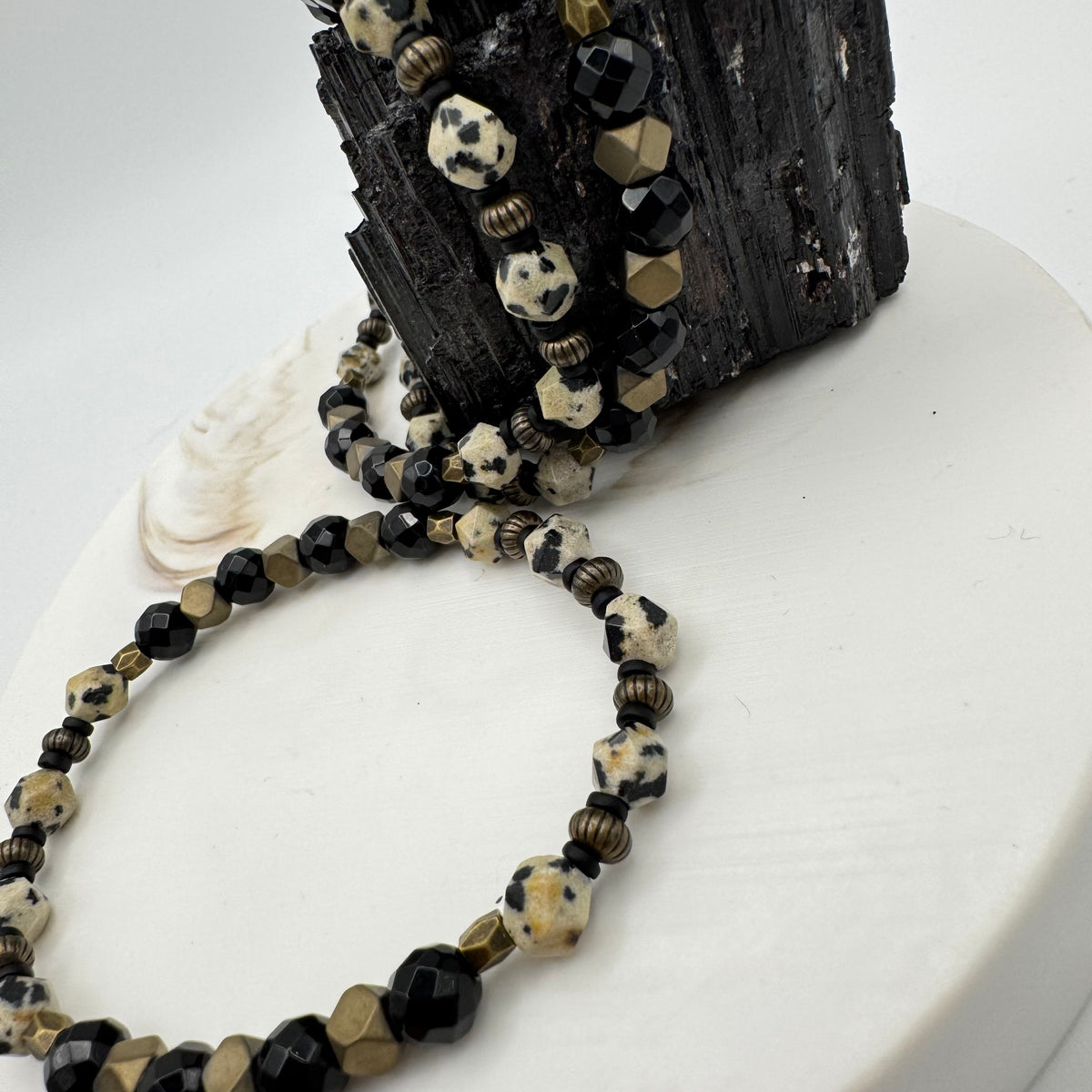 Parallel Path handmade bracelet macro close up showing faceted dalmatian jasper, black onyx and antique gold-tone ribbed and geometric beads draped on raw black tourmaline and white pedestal, detailed gemstone jewelry by Blue Jeans and Pearls Saskatchewan
