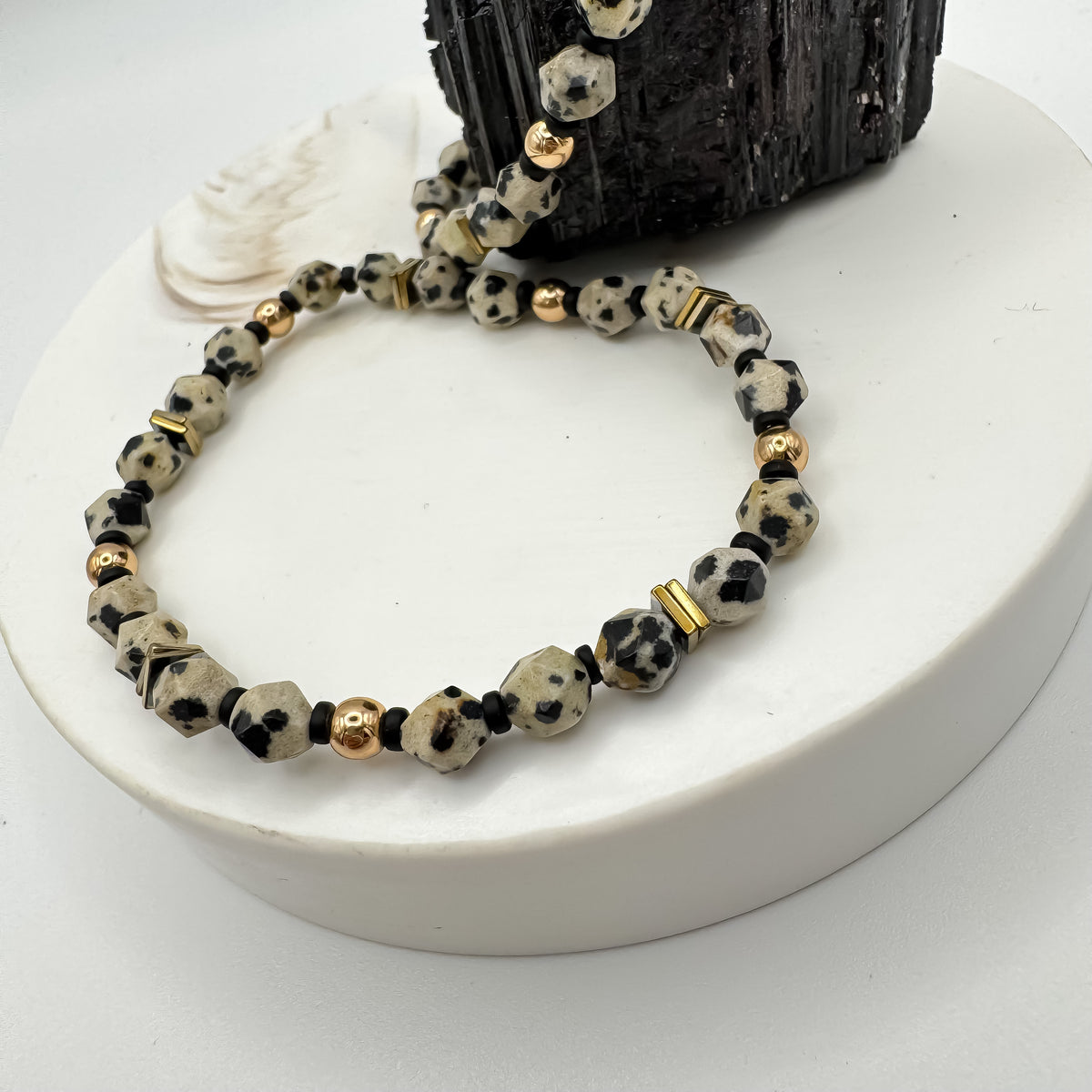 Rooted Light star cut dalmatian jasper bracelet detail with gold round beads, gold hematite square accents and black spacer beads on white pedestal with seashell and raw black tourmaline, handmade gemstone jewelry by Blue Jeans and Pearls Saskatchewan