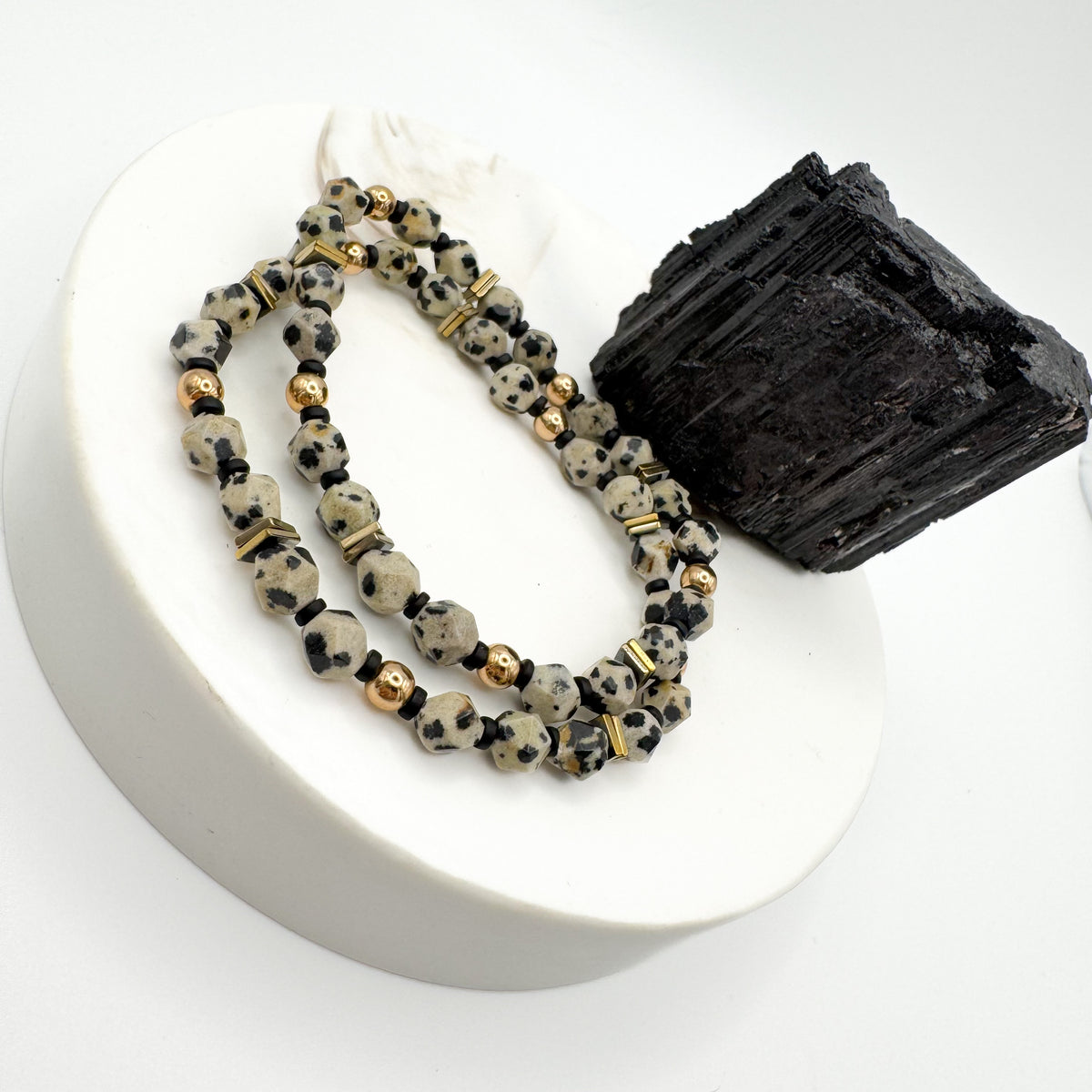 Rooted Light handmade bracelet pair with 6mm star cut dalmatian jasper, gold round beads and gold hematite square accents on white pedestal beside raw black tourmaline, stackable grounding gemstone jewelry by Blue Jeans and Pearls Saskatchewan
