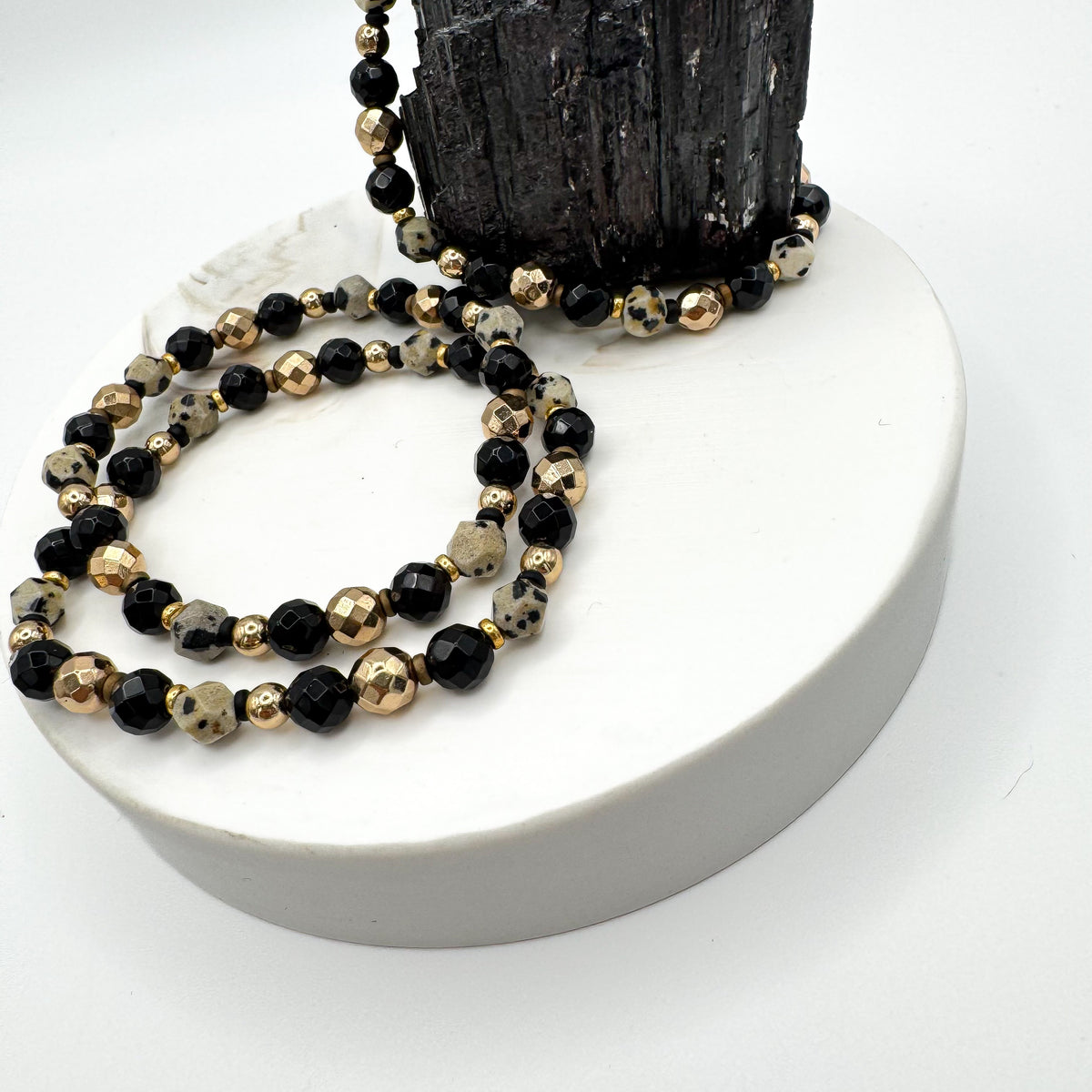 Sacred Flow handmade bracelet pair with dalmatian jasper, faceted black onyx and gold hematite beads draped on white pedestal with raw black tourmaline crystal, grounding gemstone jewelry by Blue Jeans and Pearls Saskatchewan
