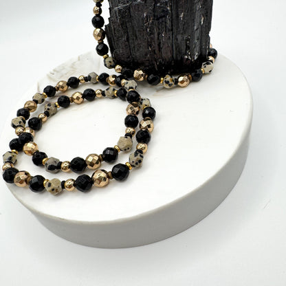 Sacred Flow handmade bracelet pair with dalmatian jasper, faceted black onyx and gold hematite beads draped on white pedestal with raw black tourmaline crystal, grounding gemstone jewelry by Blue Jeans and Pearls Saskatchewan