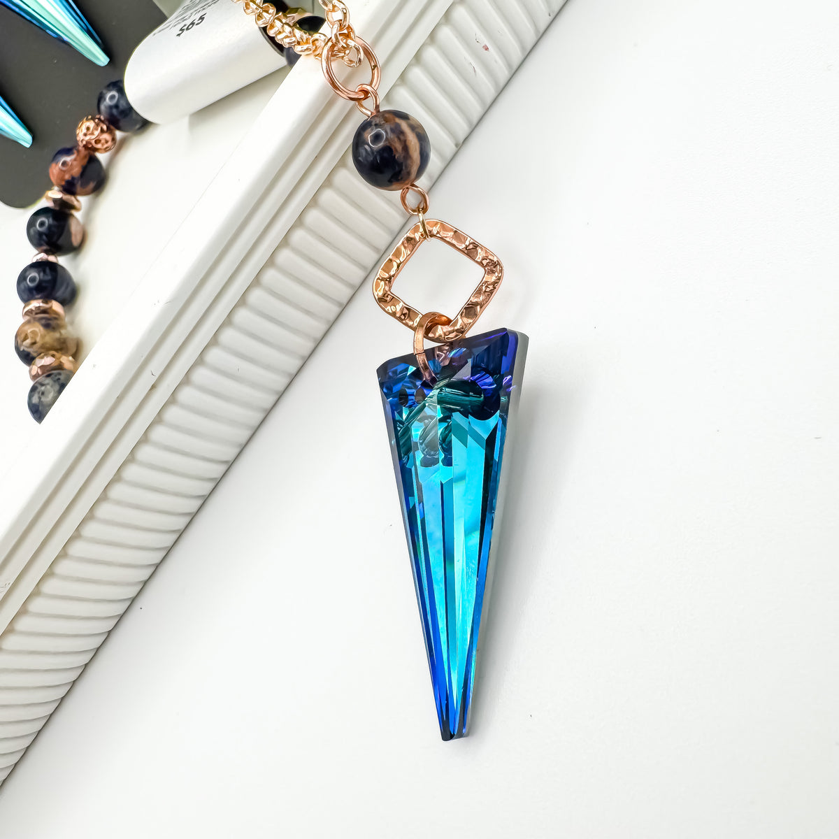 Close up of the Seas and Sunsets Necklace showing the faceted Bermuda Blue crystal spike shifting from teal to deep sapphire, with a hammered rose gold square connector and Brazilian sodalite bead with orange inclusions, by Blue Jeans and Pearls Saskatchewan