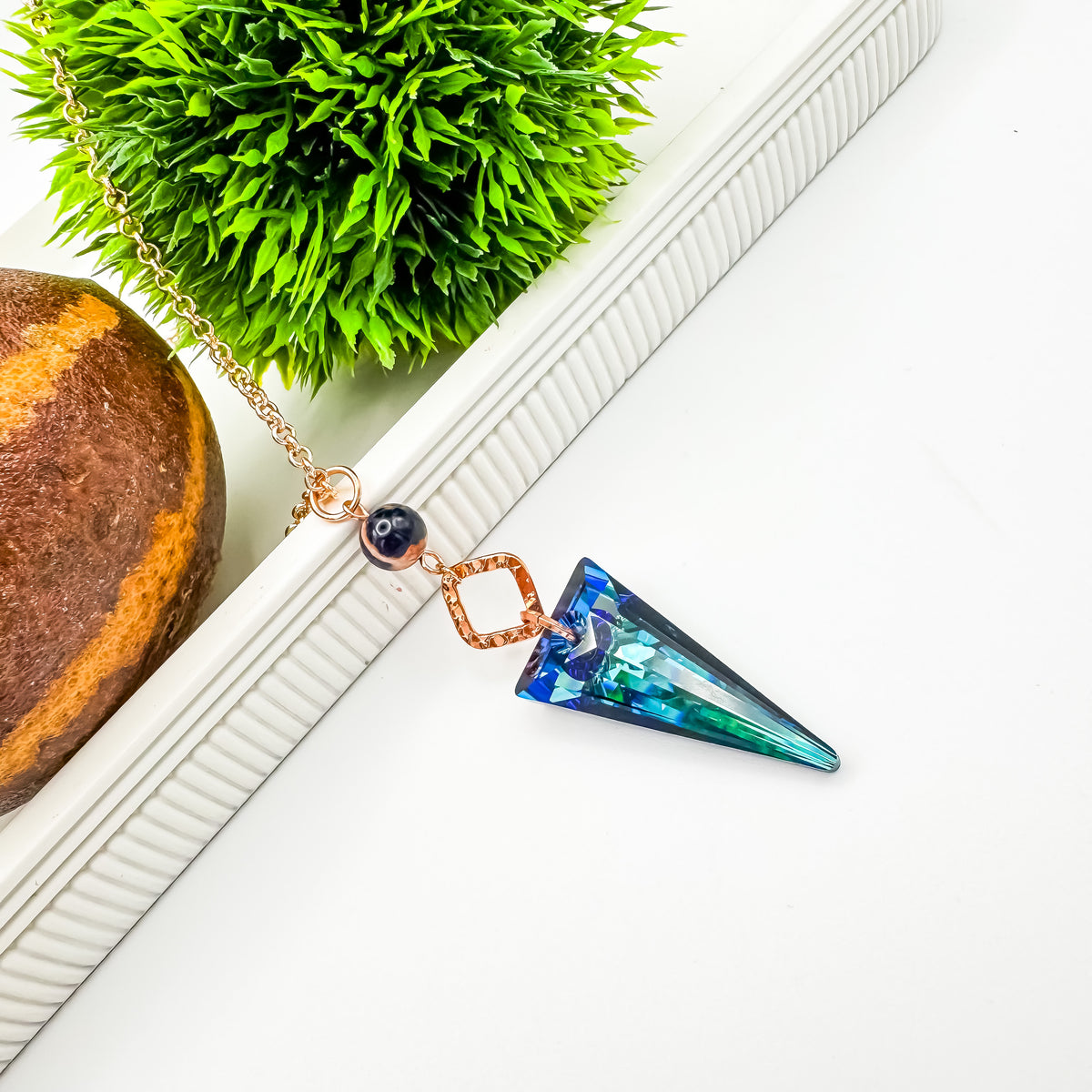 Seas and Sunsets Necklace laid flat on a white ribbed stand, the 39mm Bermuda Blue crystal spike pendant shifting from deep blue to sea green, with a Brazilian sodalite bead and hammered rose gold connector, beside greenery and a stone sphere, by Blue Jeans and Pearls Saskatchewan