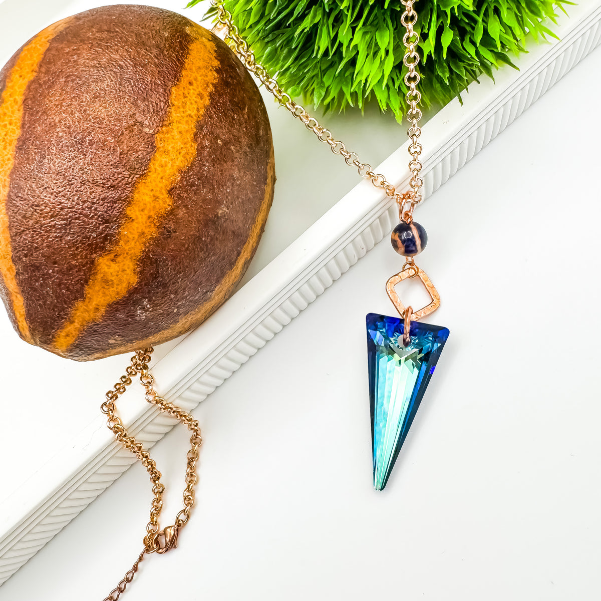 Full length view of the Seas and Sunsets Necklace showing the rose gold plated rolo chain and lobster clasp draped around a striped stone sphere, with the Bermuda Blue crystal spike pendant and Brazilian sodalite bead, by Blue Jeans and Pearls Saskatchewan