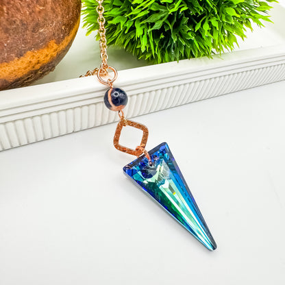 Seas and Sunsets Necklace draped over a white ribbed stand, showing the Bermuda Blue crystal spike pendant in blue and sea green, a Brazilian sodalite bead with an orange sunset swirl, and a hammered rose gold square connector on a rose gold chain, by Blue Jeans and Pearls Saskatchewan