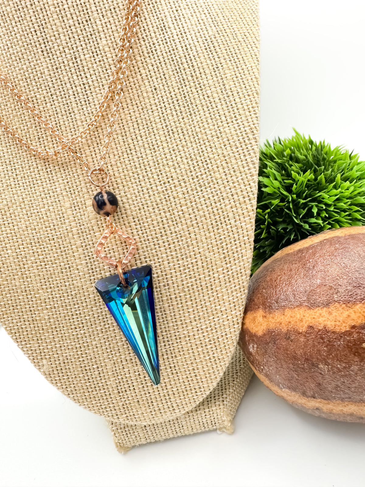 Side angle of the Seas and Sunsets Necklace on a burlap jewelry bust, Bermuda Blue crystal spike pendant hanging below a Brazilian sodalite bead and rose gold connector, styled with greenery and a striped stone sphere, by Blue Jeans and Pearls Saskatchewan