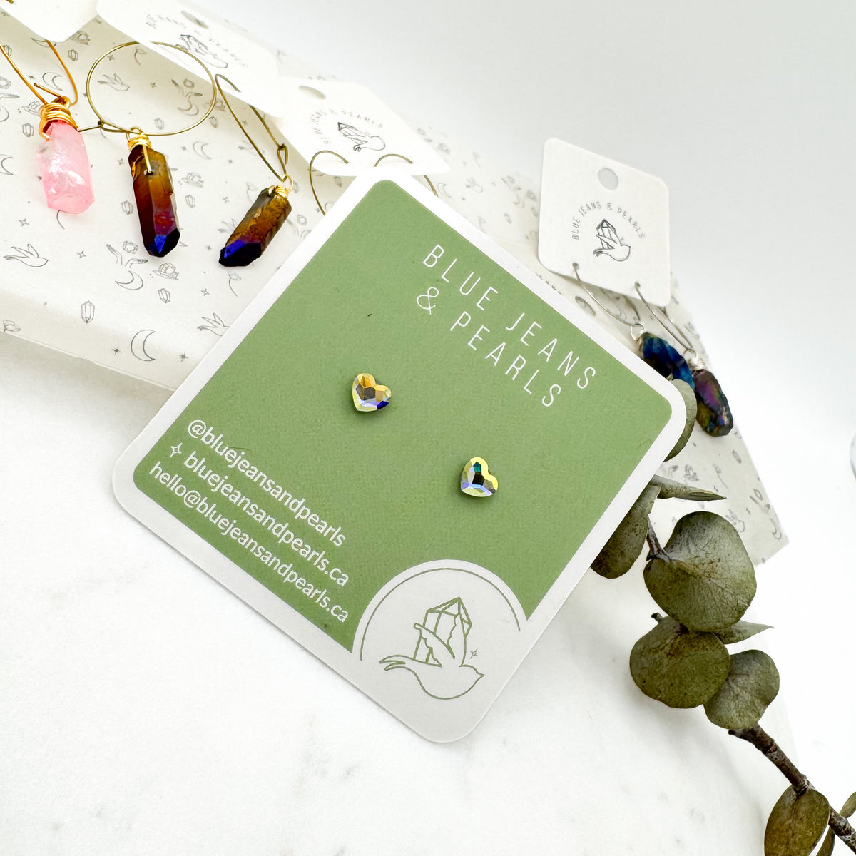 Small Heart Stud earrings in aurora borealis Austrian crystal on a sage green Blue Jeans and Pearls card, styled with aura crystal point hoop earrings and eucalyptus, metal-free studs for sensitive ears by Blue Jeans and Pearls Saskatchewan