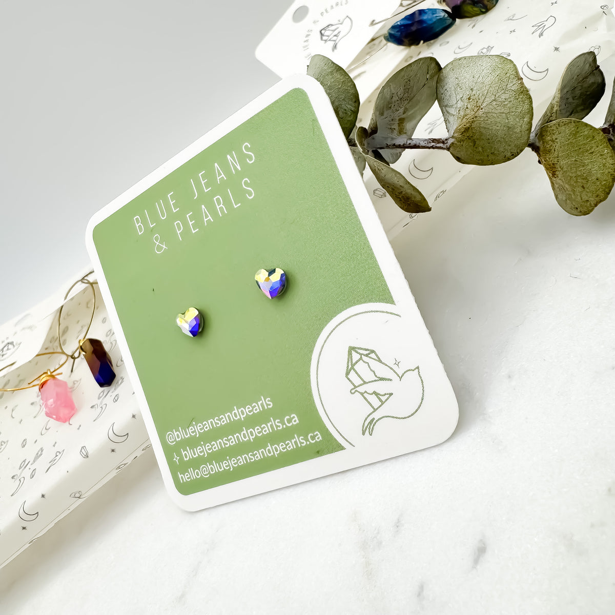 Close up of Small Heart Stud earrings in aurora borealis Austrian crystal on an upright sage green Blue Jeans and Pearls card, rainbow shimmer catching the light with eucalyptus and crystal earrings behind, metal-free studs by Blue Jeans and Pearls Saskatchewan