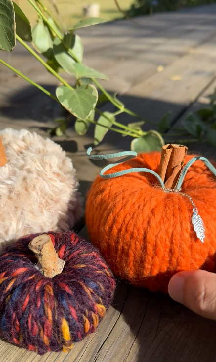 Three handmade yarn pumpkins on a sunny wooden deck: a bright orange chunky knit pumpkin with a cinnamon stick stem and silver leaf charm, a fluffy cream pumpkin, and a deep plum multicolour pumpkin with a wood stem, from the DIY yarn pumpkin workshop