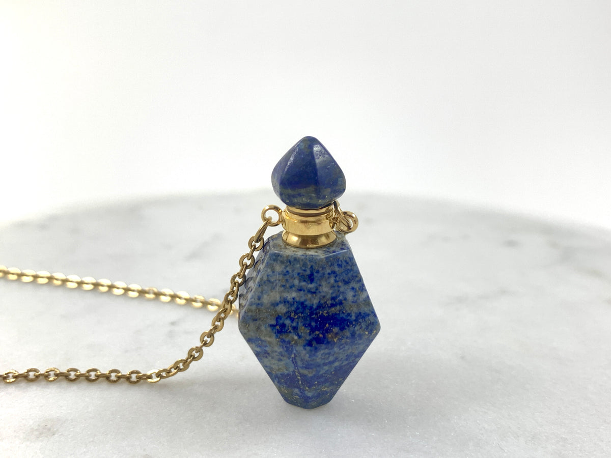 Gold Lapis Lazuli Love Potion Bottle Necklace with a faceted deep blue lapis lazuli crystal bottle pendant, gold plated cap and stopper on a gold chain, displayed on white marble, essential oil and perfume vial necklace by Blue Jeans and Pearls Saskatchewan