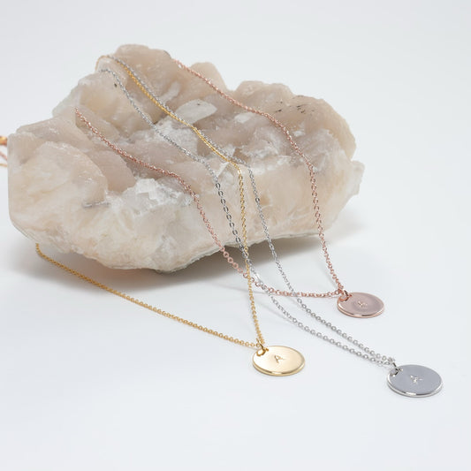 Hand Stamped Initial Disc Add On options shown in yellow gold, white gold and rose gold plated 11mm round discs stamped with the letter A on matching chains, draped over a raw pale pink crystal cluster, by Blue Jeans and Pearls Saskatchewan