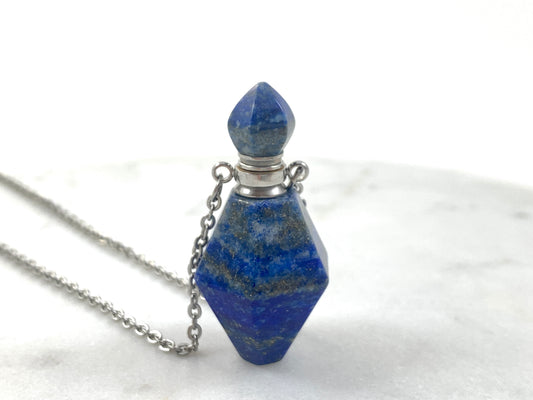 Silver Lapis Lazuli Love Potion Bottle Necklace with a faceted deep blue lapis lazuli crystal bottle pendant flecked with gold pyrite, silver tone cap and stopper on a silver chain, displayed on white marble, essential oil and perfume vial necklace by Blue Jeans and Pearls Saskatchewan