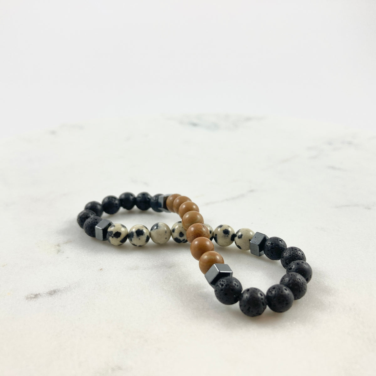Men's handmade dalmatian jasper and wood jasper bracelet pair with lava stone and hematite cube accents on marble surface, natural earthy diffuser jewelry by Blue Jeans and Pearls Regina Saskatchewan